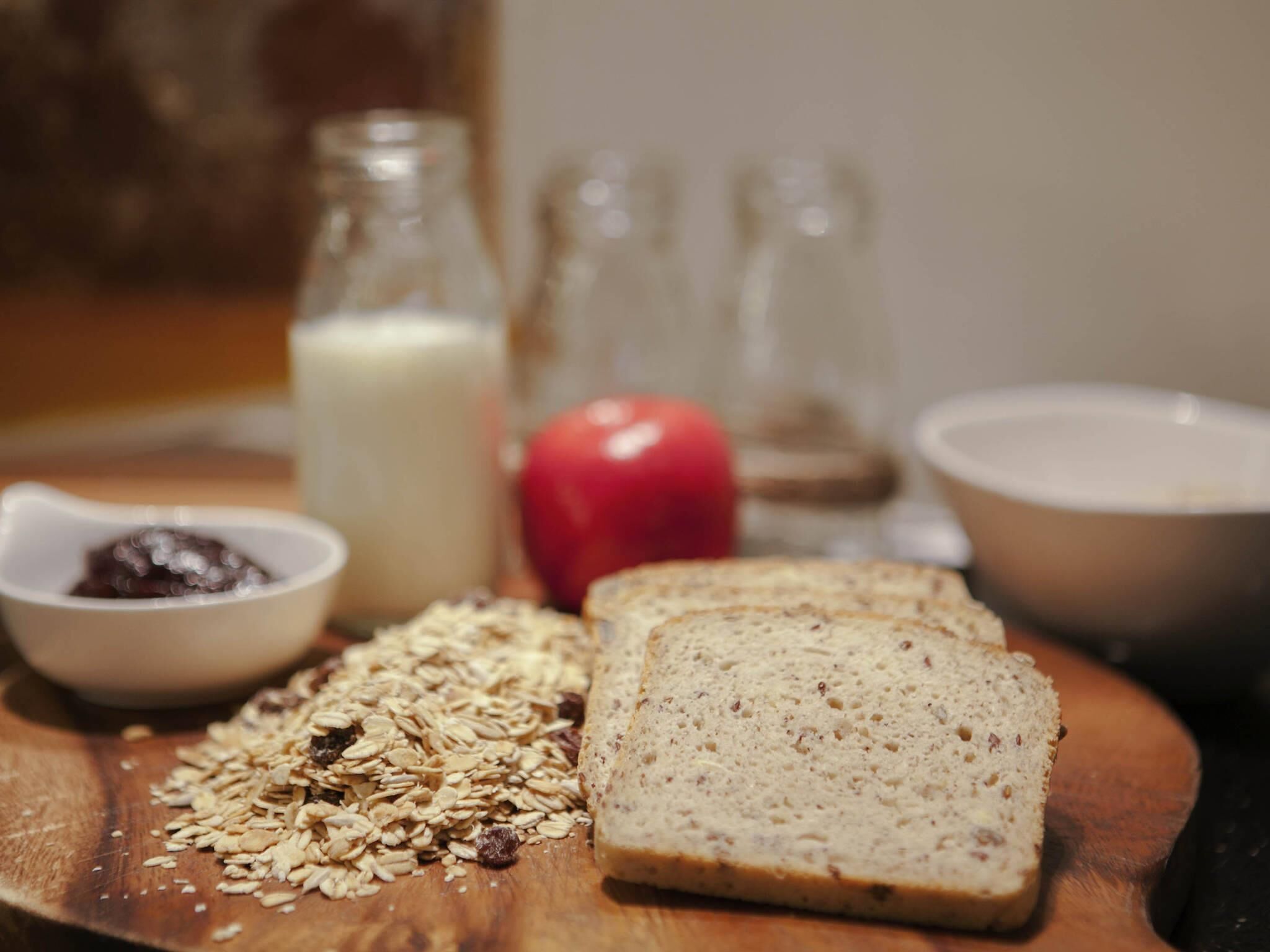 A wooden cutting board with sliced bread, oats, a red apple, a glass bottle of milk, and a small bowl of jam.