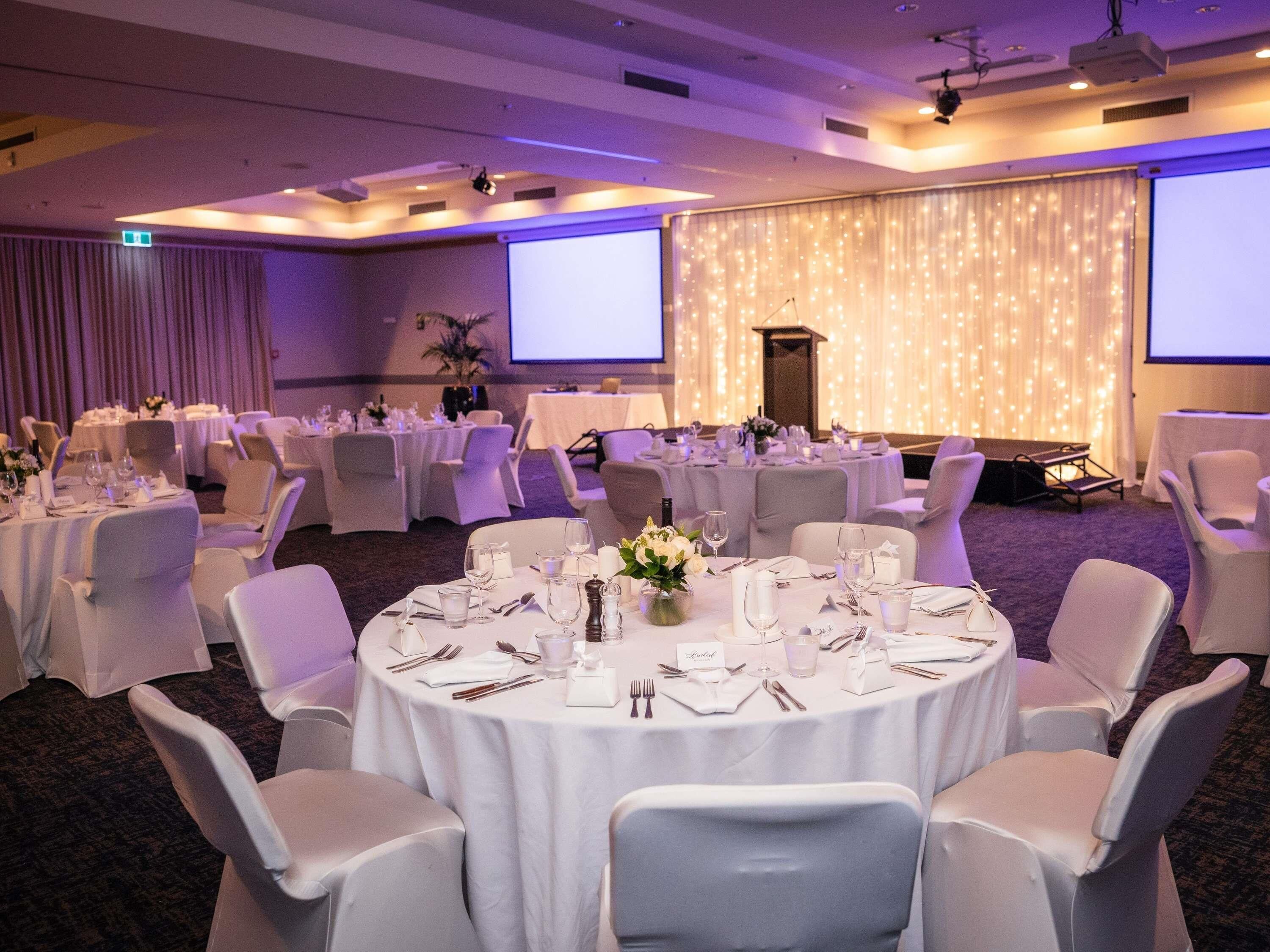 Une salle de banquet avec des tables rondes dressées pour le repas, des chaises blanches et une scène équipée d’un écran.