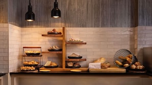 A breakfast buffet with various breads and pastries, wooden shelves, and black pendant lights.
