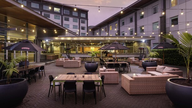 An outdoor patio area with tables, chairs, and umbrellas, surrounded by buildings with multiple windows and string lights.