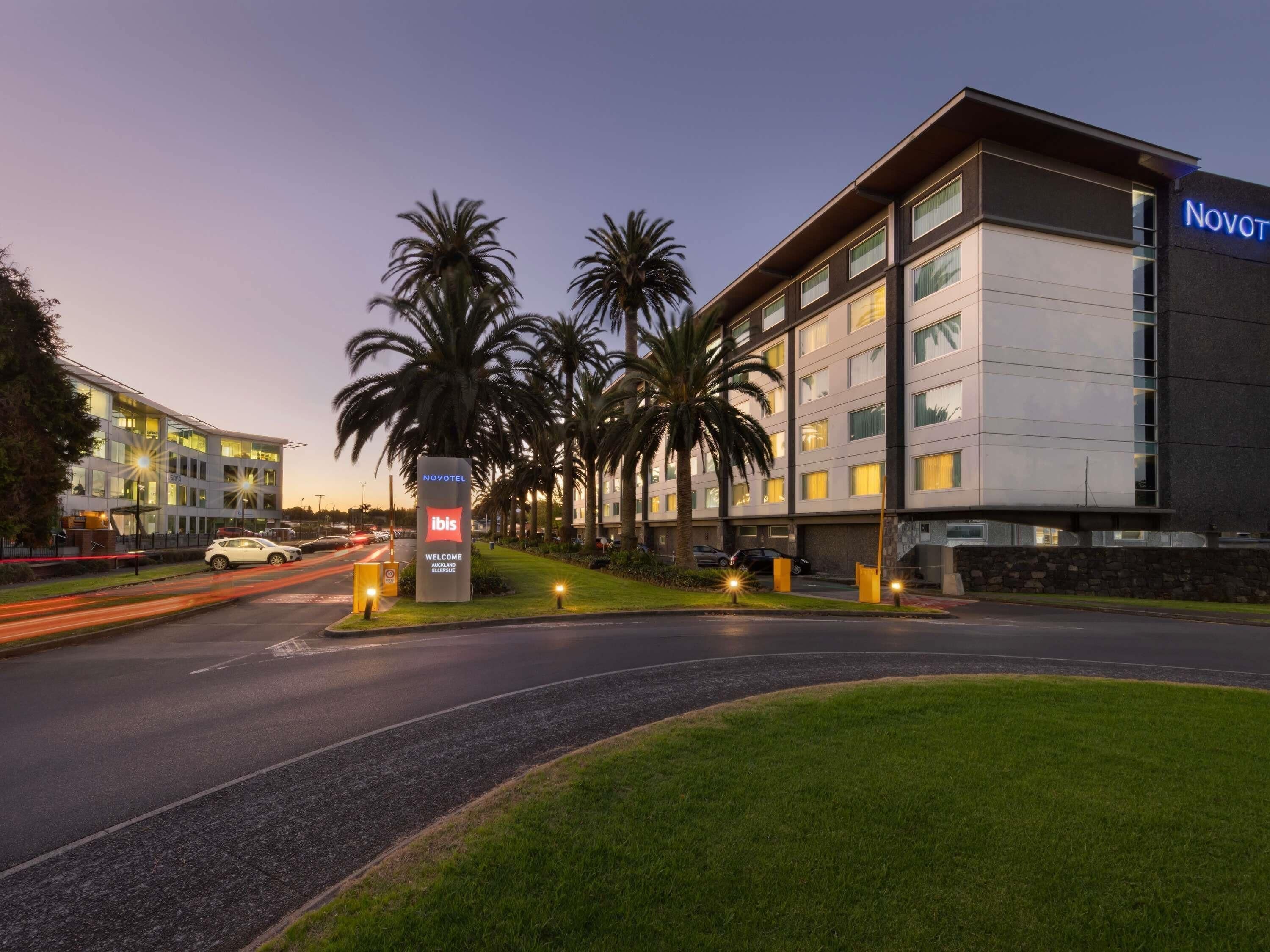 A modern hotel with a signboard, palm trees, and a well-lit entrance.