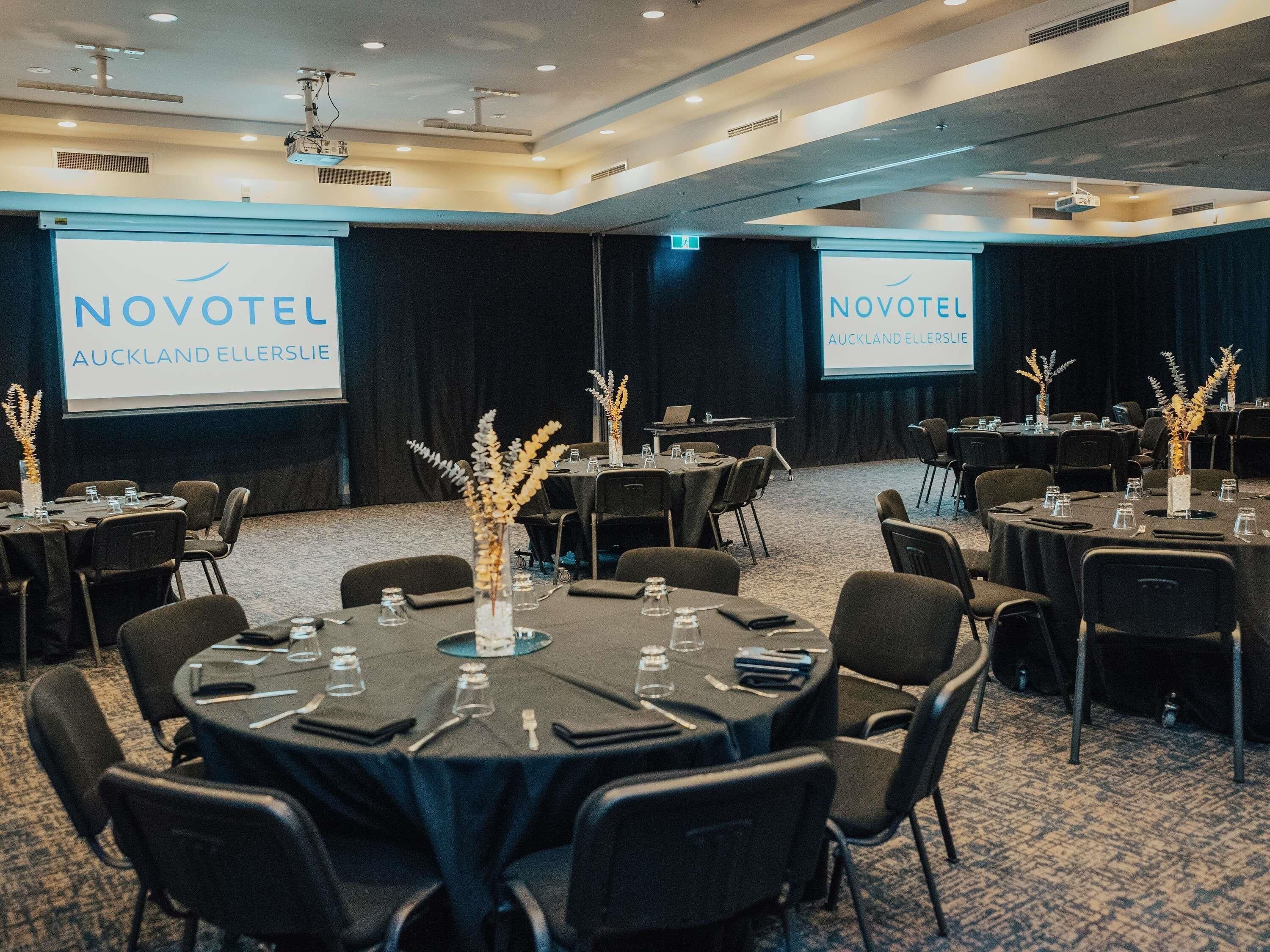 A conference room with round tables, chairs, and a projector screen displaying 'NOVOTEL AUCKLAND ELLERSLIE'.