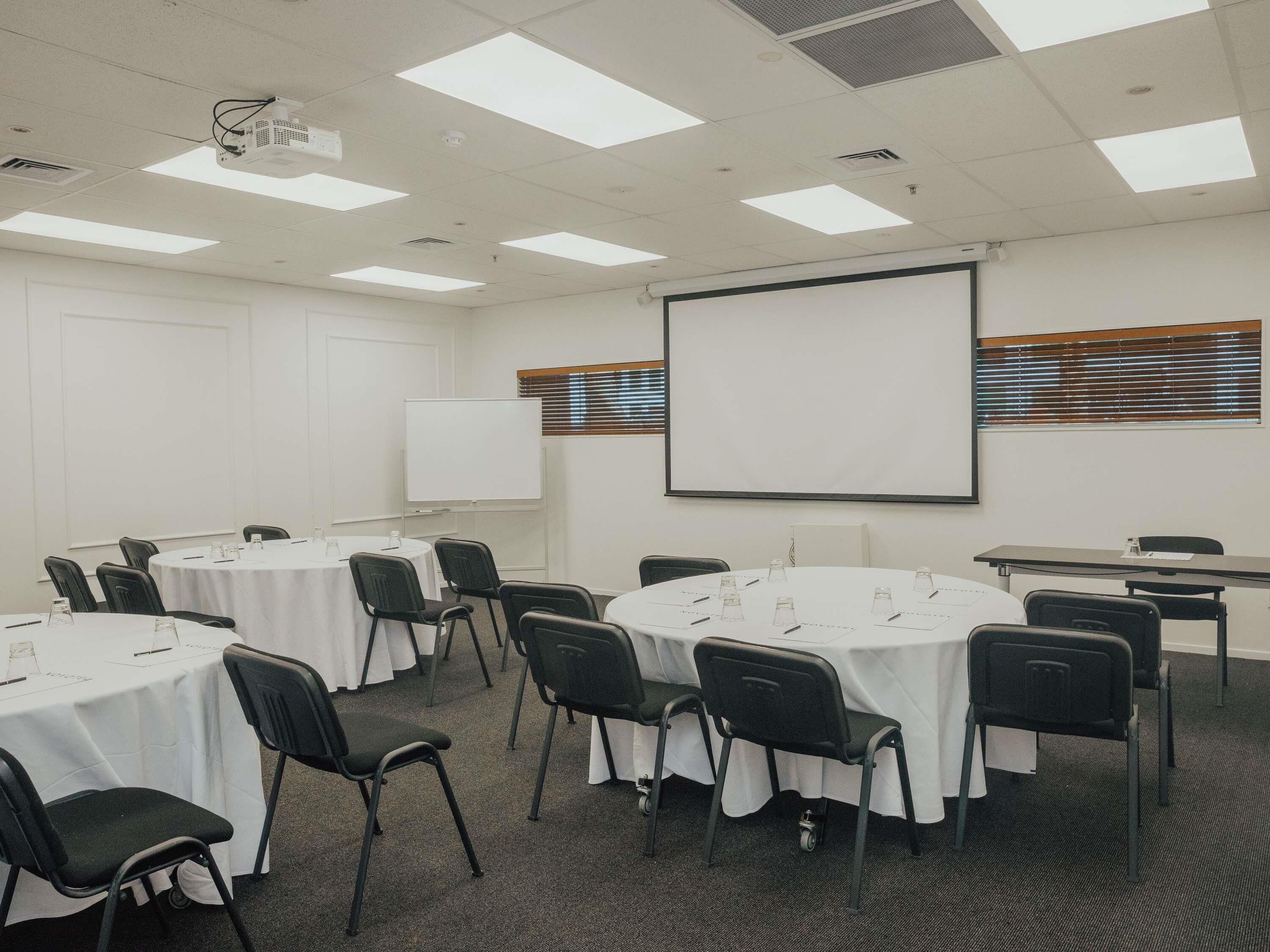 A conference room with round tables, black chairs, a whiteboard, a projector, and a large screen.