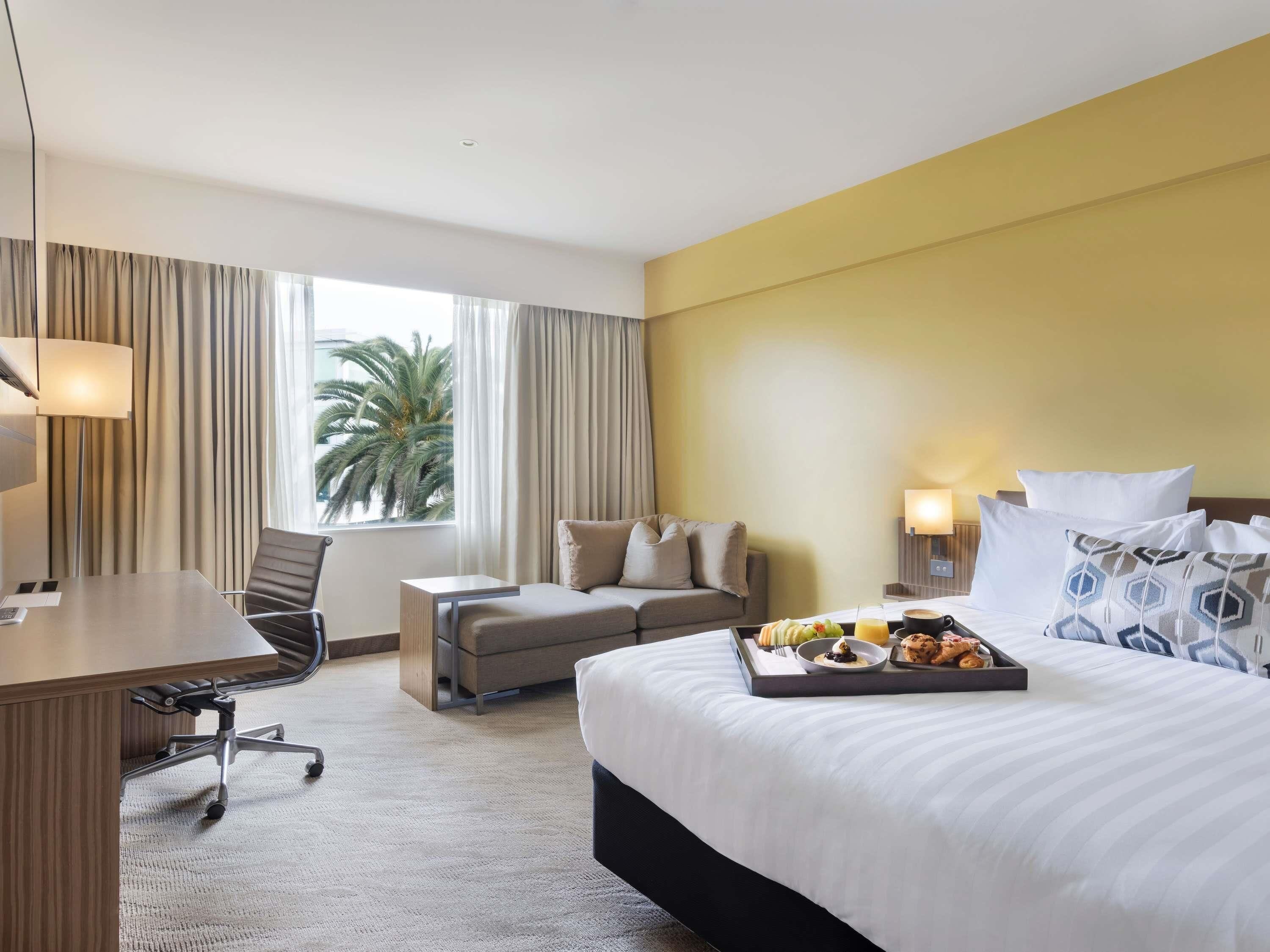 Une chambre d’hôtel comprenant un lit, un bureau, une chaise, un canapé, une fenêtre avec des rideaux et un plateau avec des articles pour le petit-déjeuner.
