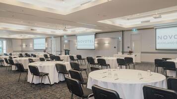 Un salón de conferencias con varias mesas y sillas, una pantalla de proyector y una pantalla de presentación que muestra 'NOVOTEL AUCKLAND ELLERSLIE'.
