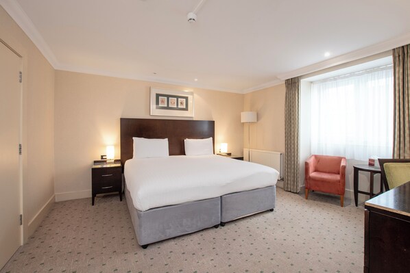 A hotel room with a large bed, bedside tables, a desk, a chair, a window with curtains, and a framed picture on the wall.