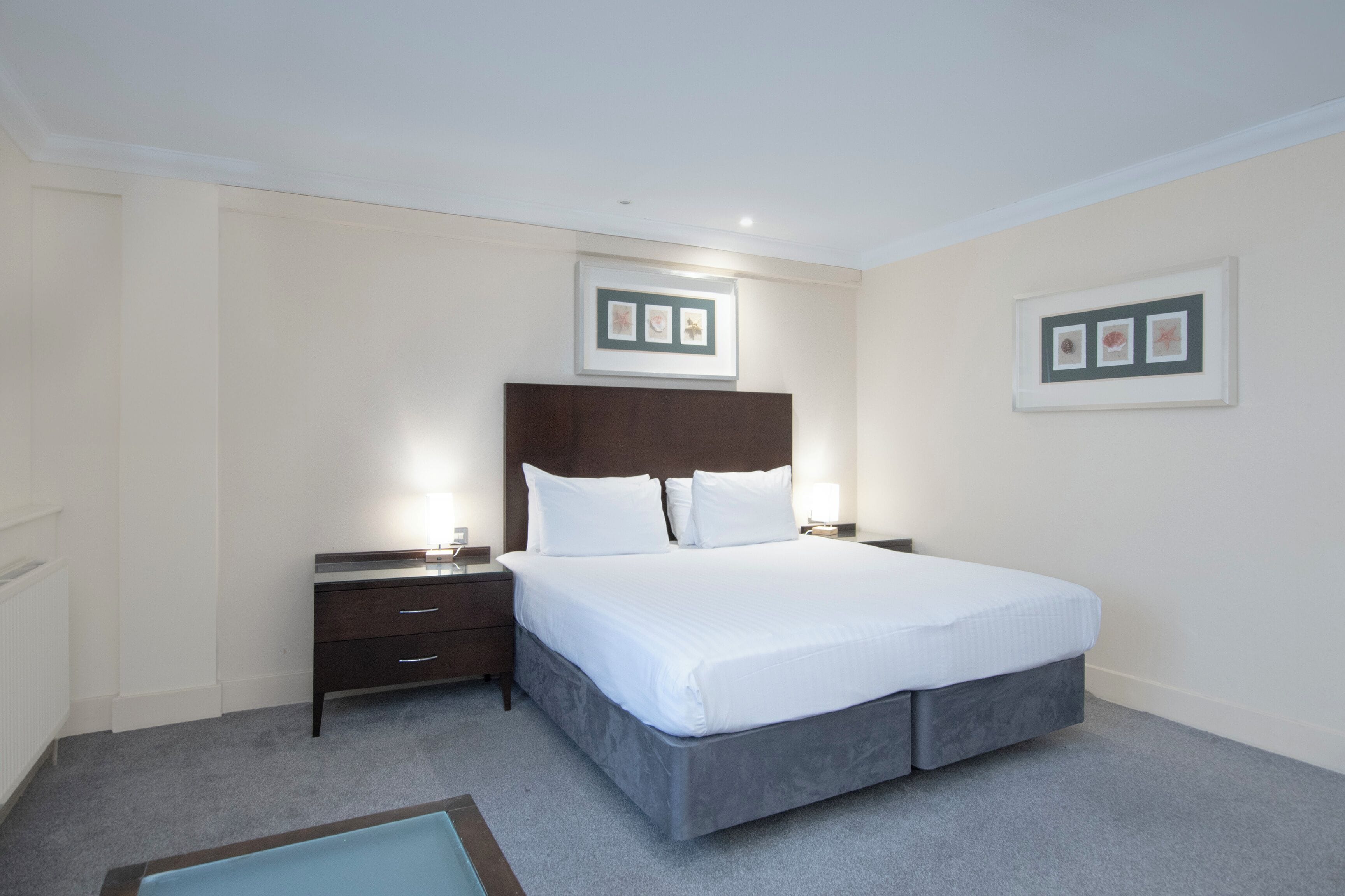 Una habitación de hotel con cama, mesita de noche, cómoda y cuadros en la pared.