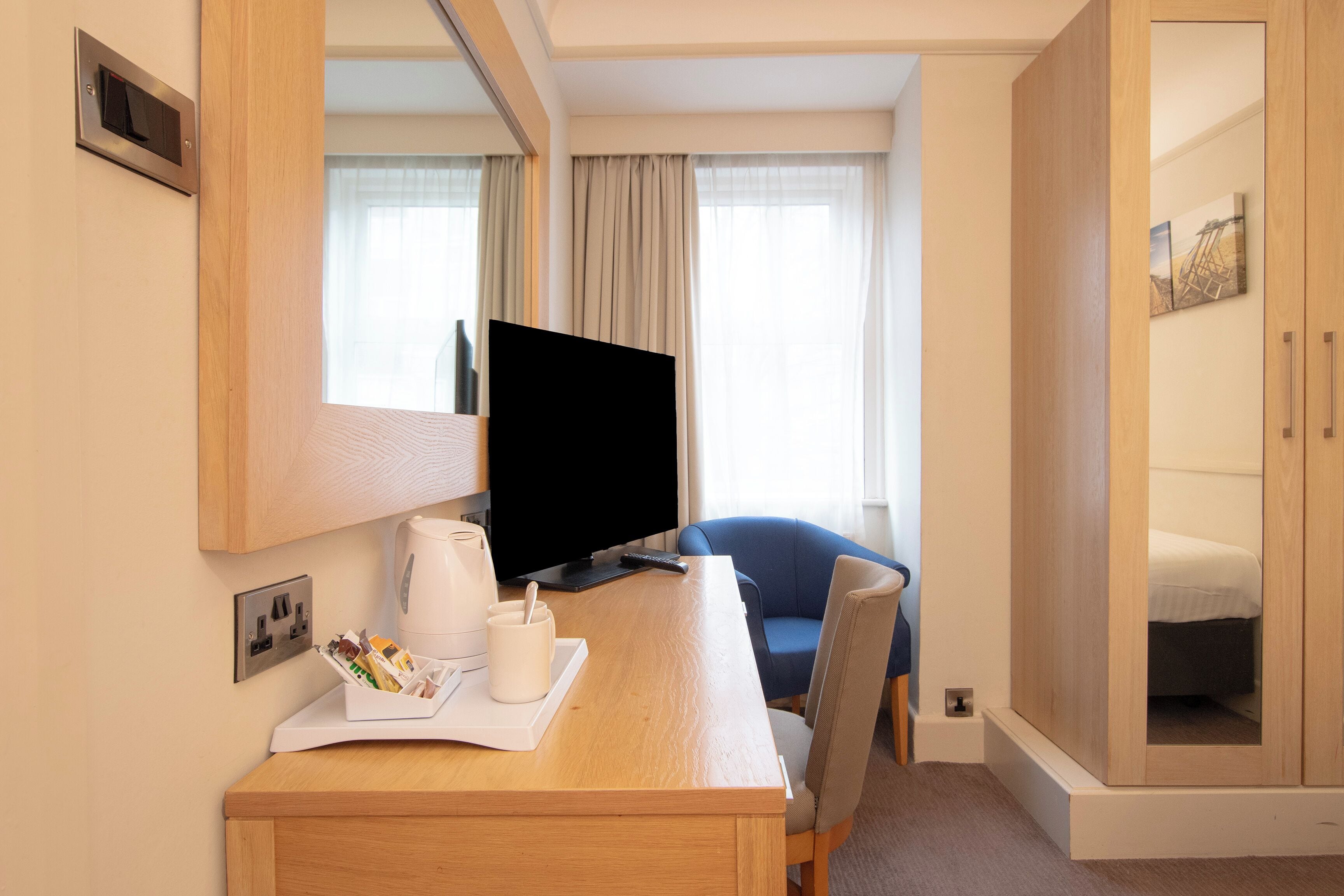 A hotel room with a wooden desk, a flat-screen TV, a chair, a bed, and a wardrobe.