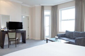 Ein Hotelzimmer mit Bett, Schreibtisch mit Fernseher, Sessel, Sofa und einem Fenster mit Vorhängen.