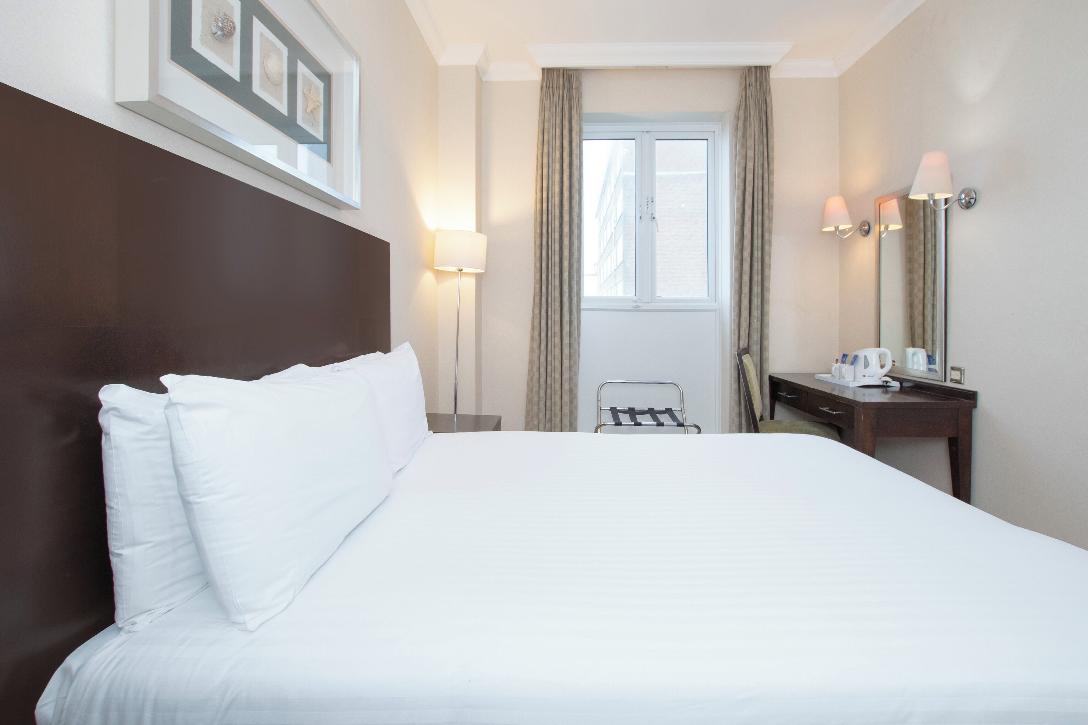 A hotel room with a large bed, a bedside table with a lamp, a desk with a mirror, and a window with curtains.