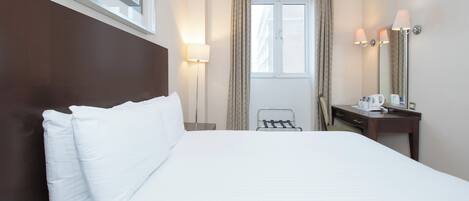 A hotel room with a large bed, a bedside table with a lamp, a desk with a mirror, and a window with curtains.