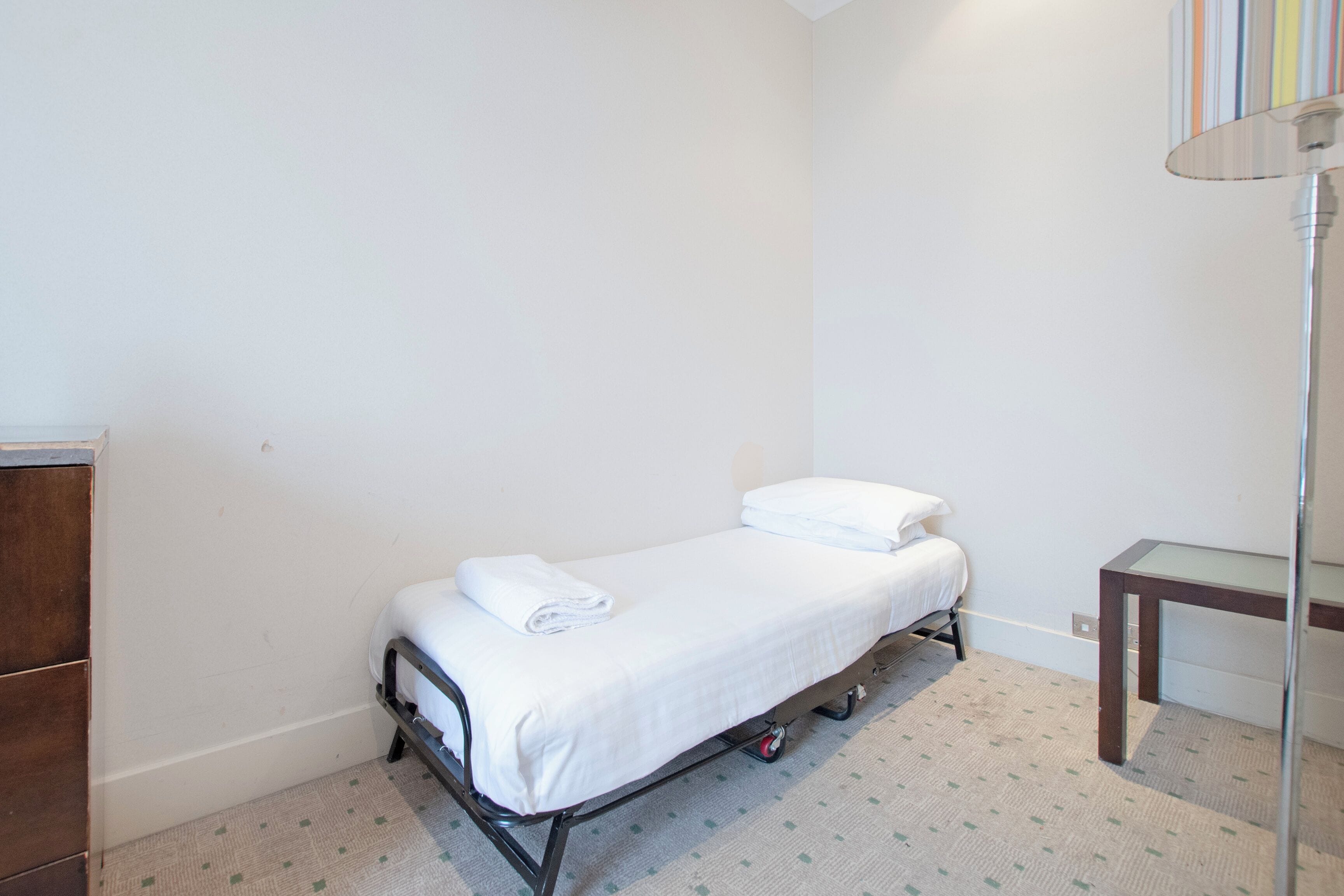 Una habitación pequeña con una cama individual, un tocador de madera, una mesita y una lámpara.