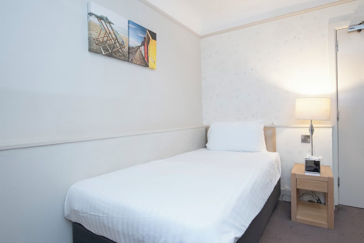 Un lit simple avec une couette blanche, une table de chevet avec une lampe et un tableau accroché au mur.
