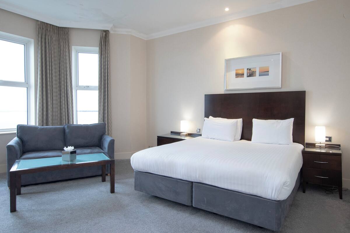 Une chambre d’hôtel avec un lit, un canapé, une table de chevet, une lampe et un tableau au mur.