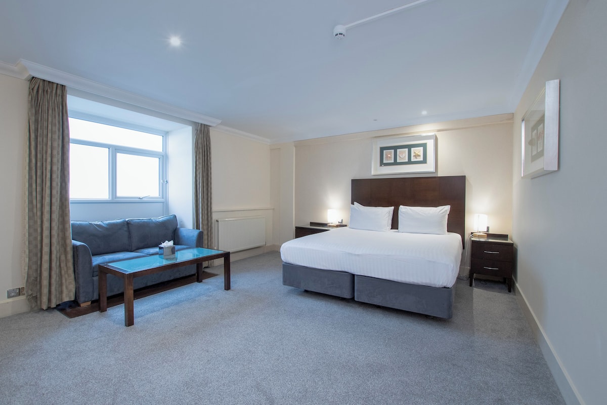 Une chambre d’hôtel avec un lit, un canapé, une petite table, une table de chevet et une fenêtre avec des rideaux.