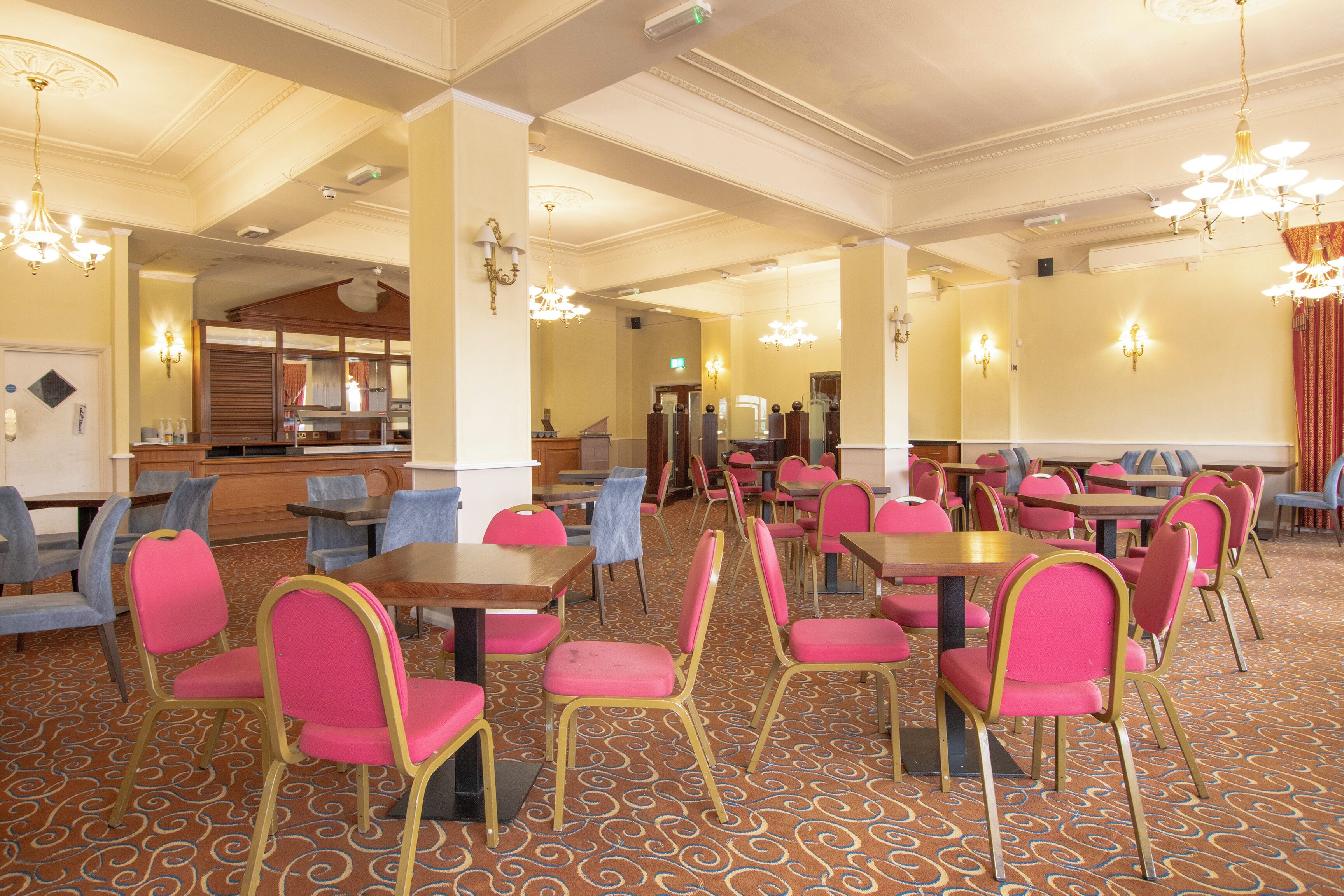 A hotel dining area with tables and chairs, a patterned carpet, and chandeliers.