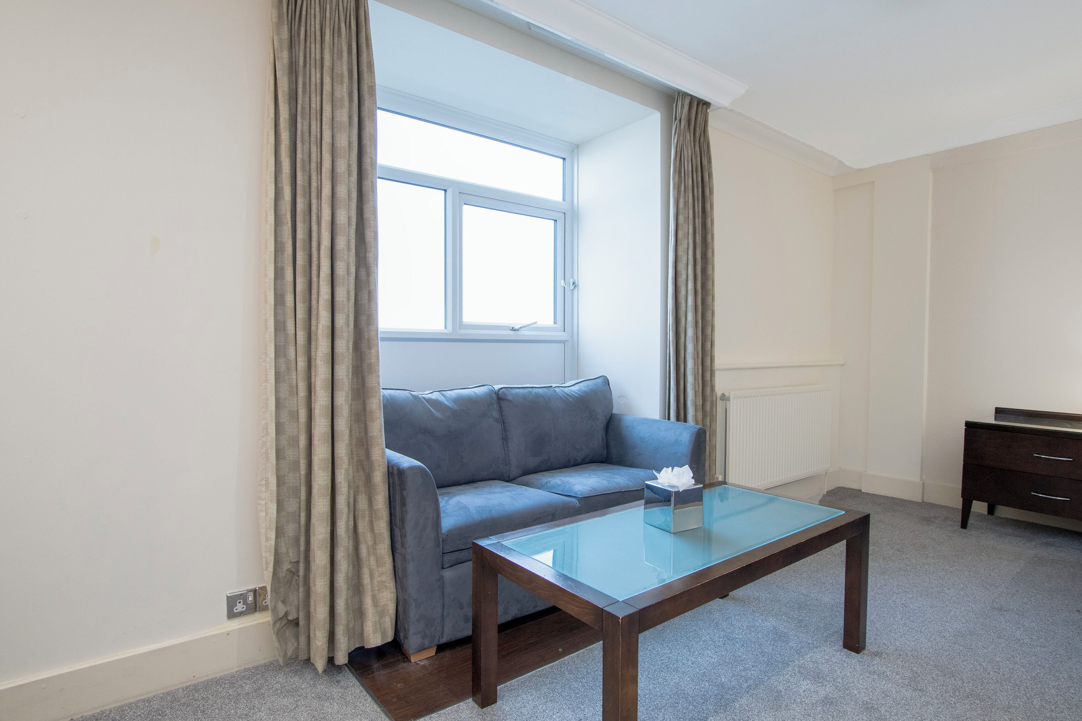 Ein Zimmer mit einem blauen Sofa, einem Glastisch, einem Heizkörper und einem Fenster mit Vorhängen.