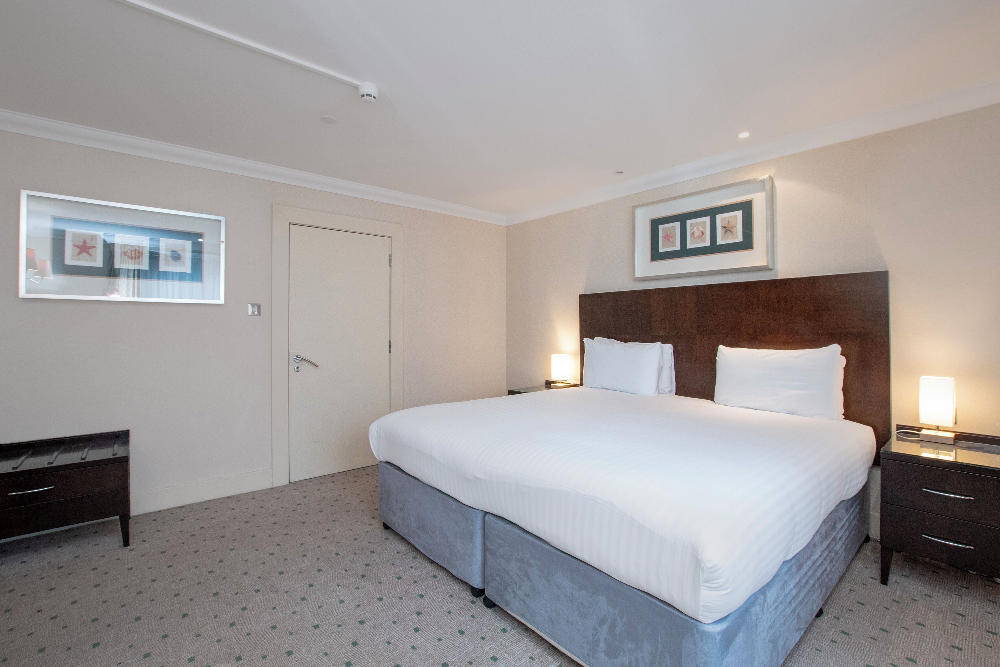 A hotel room with a large bed, a bedside table, a wardrobe, and framed pictures on the wall.