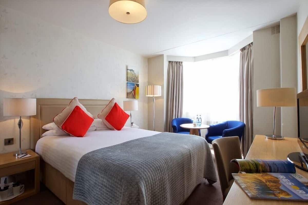 Une chambre d’hôtel avec un lit, un bureau, une chaise, une lampe et une fenêtre.