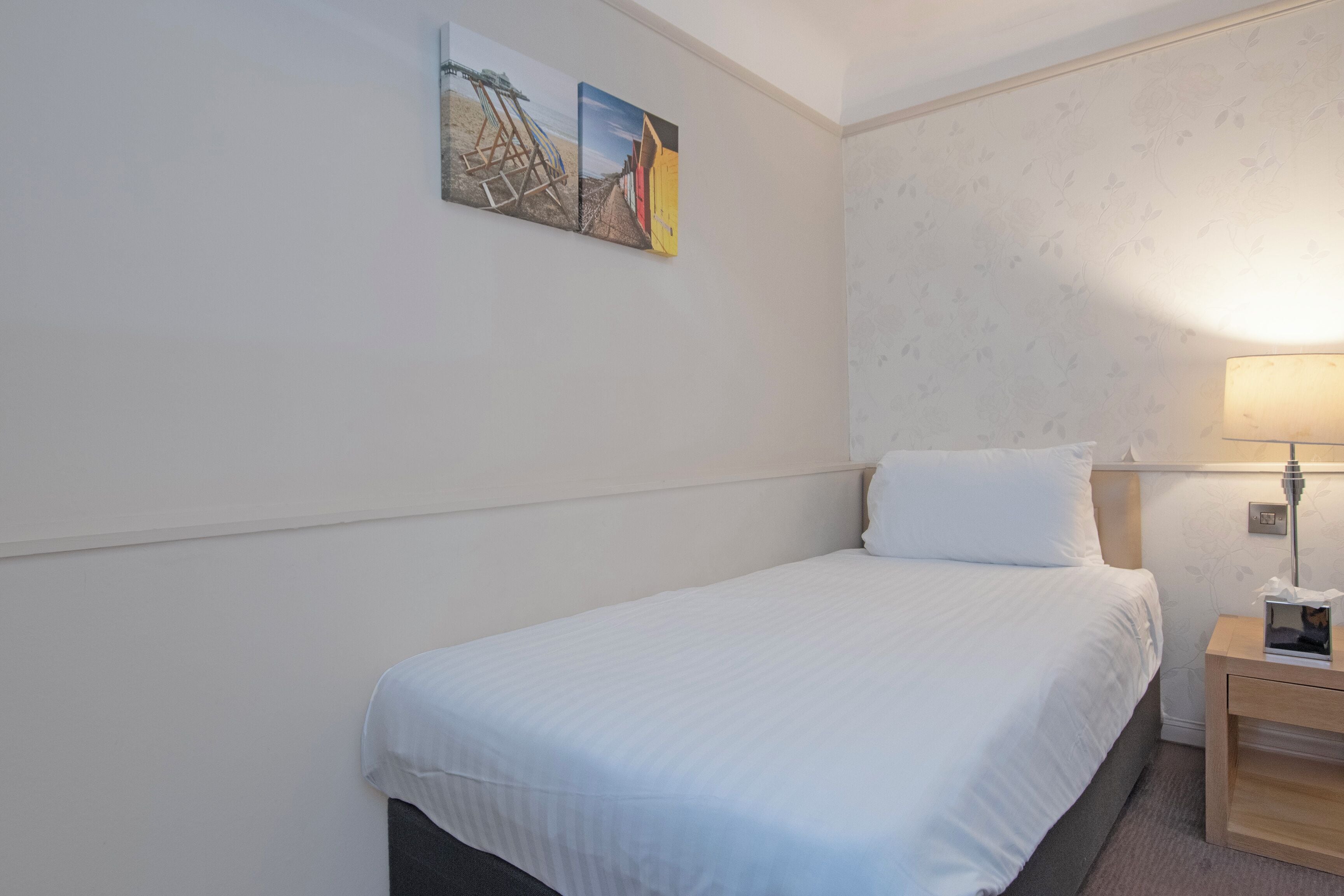 A hotel room with a single bed, a nightstand with a lamp, and a wall-mounted painting.