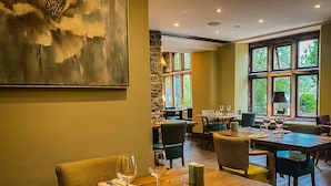 Un restaurante con mesas y sillas de madera, un cuadro en la pared y grandes ventanales con vistas a zonas verdes.