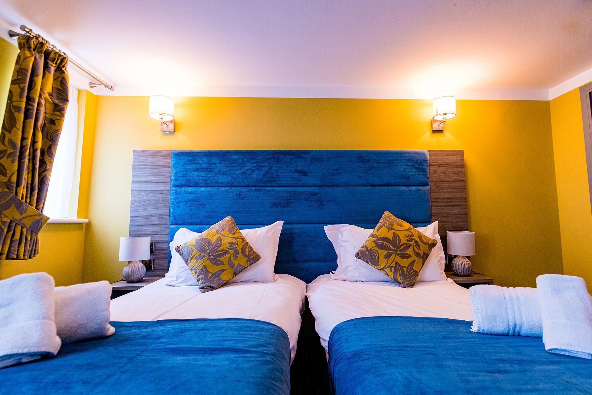 A hotel room with two beds, each with a blue headboard and white bedding. There are two wall-mounted lamps on either side of the bed, and a window with yellow curtains.