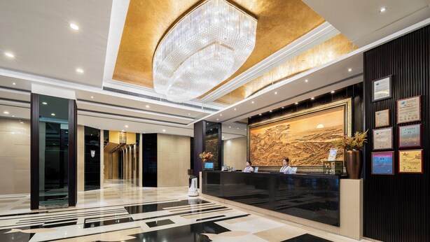 Een moderne hotellobby met een receptiebalie, een grote kroonluchter en een marmeren wand met certificaten.