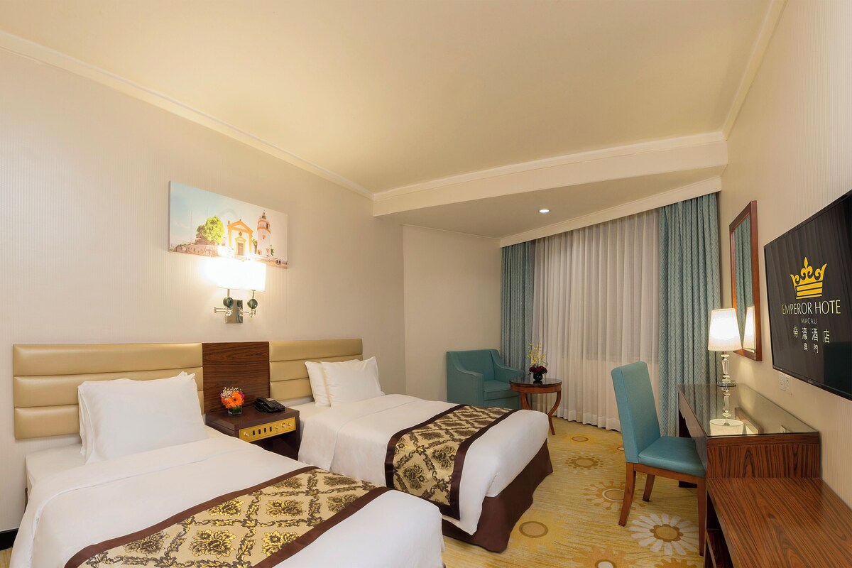 A hotel room with two beds, a desk, a chair, a nightstand, a lamp, a painting, and a curtain.