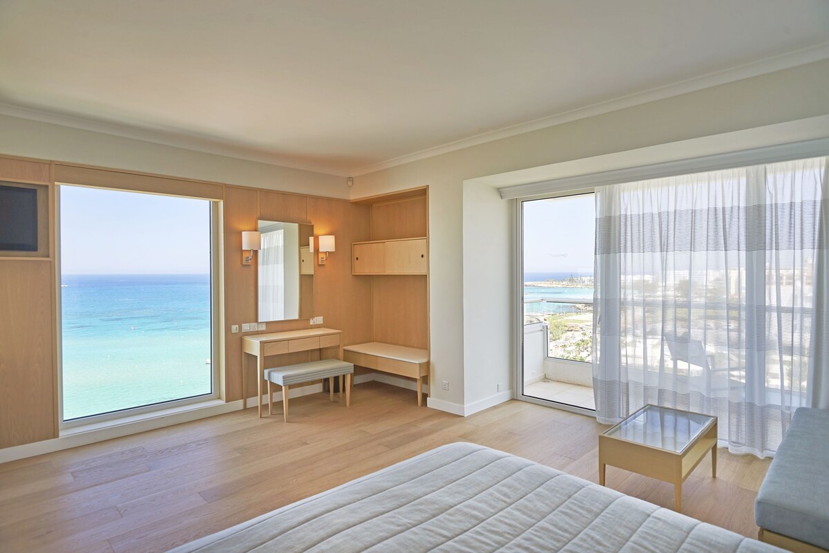 A hotel room with a large window, a bed, a desk, a bench, and a chair.