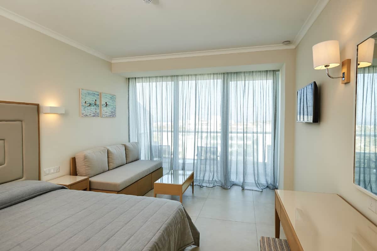Une chambre d’hôtel avec un lit, un canapé, une petite table, une télévision et une fenêtre avec des rideaux légers.