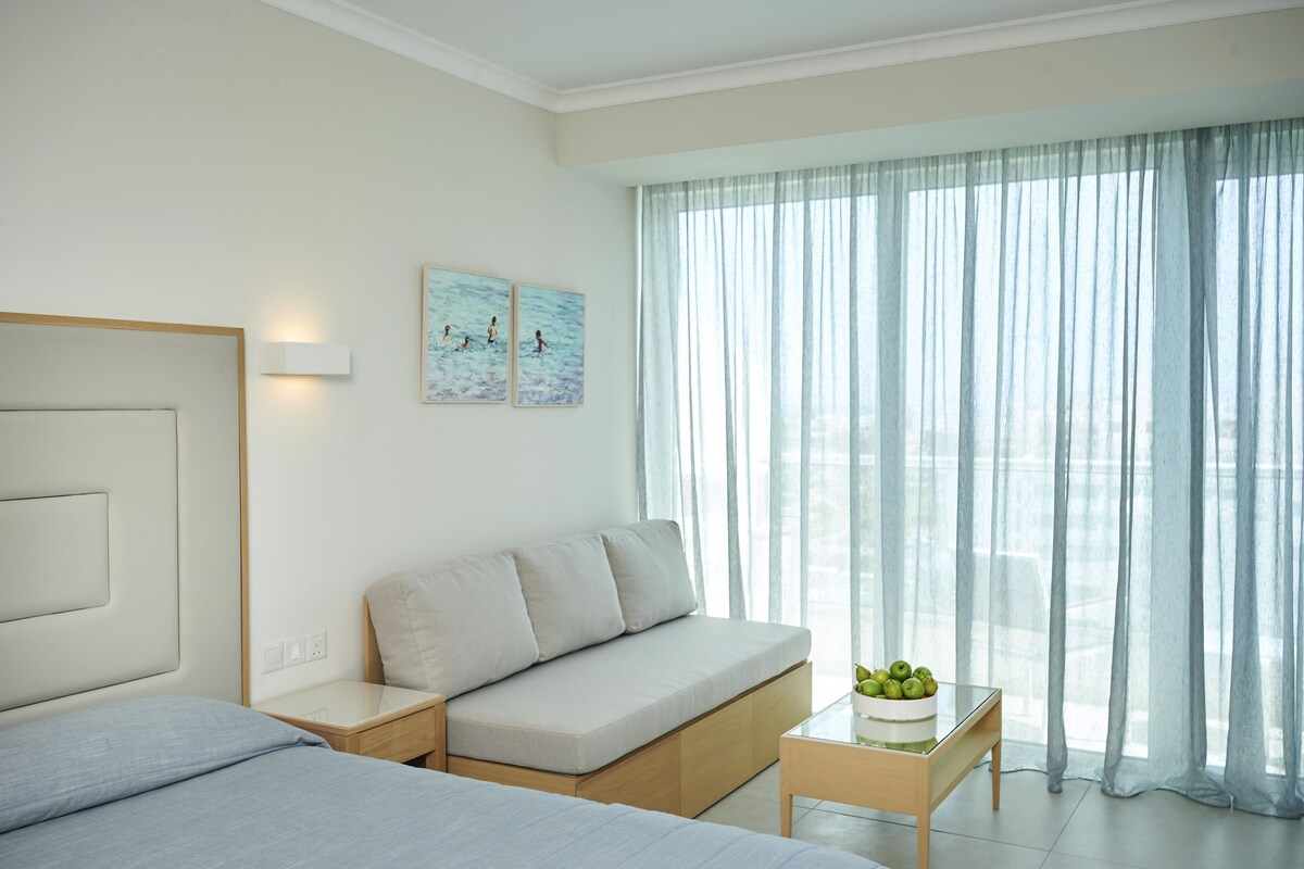 A modern hotel room with a bed, a sofa, a small table with fruit, and a large window with sheer curtains.
