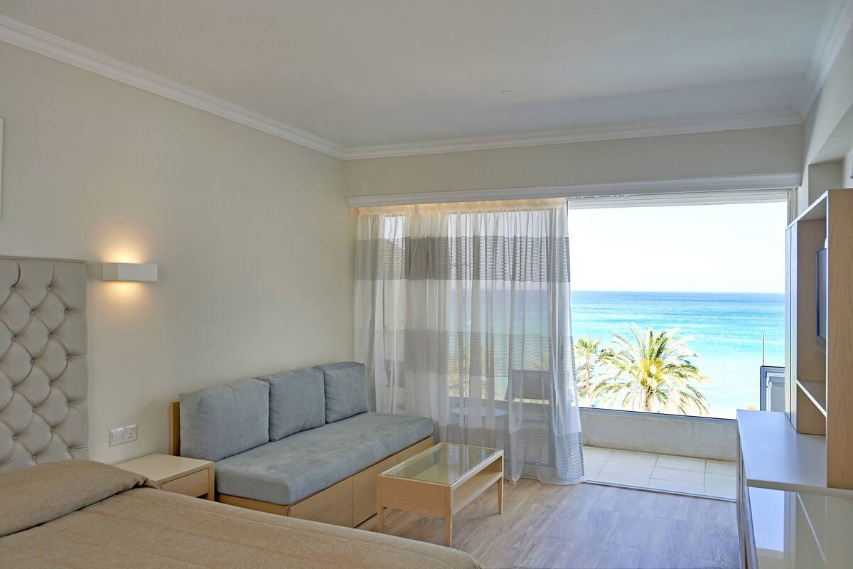 Une chambre moderne avec un canapé, une table basse en verre, un téléviseur et un balcon offrant une vue sur l’océan.