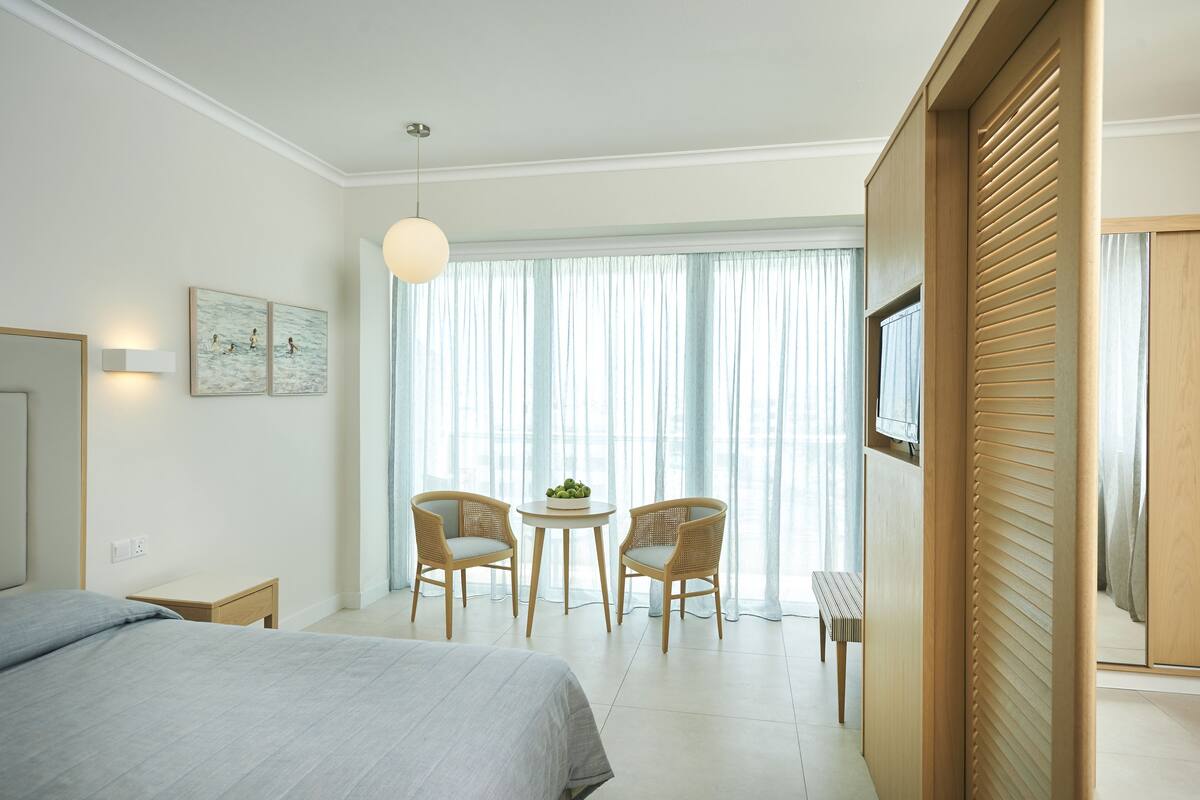 Une chambre d’hôtel avec un lit, deux fauteuils en osier, une petite table avec une plante, un banc et une grande fenêtre avec des rideaux légers.