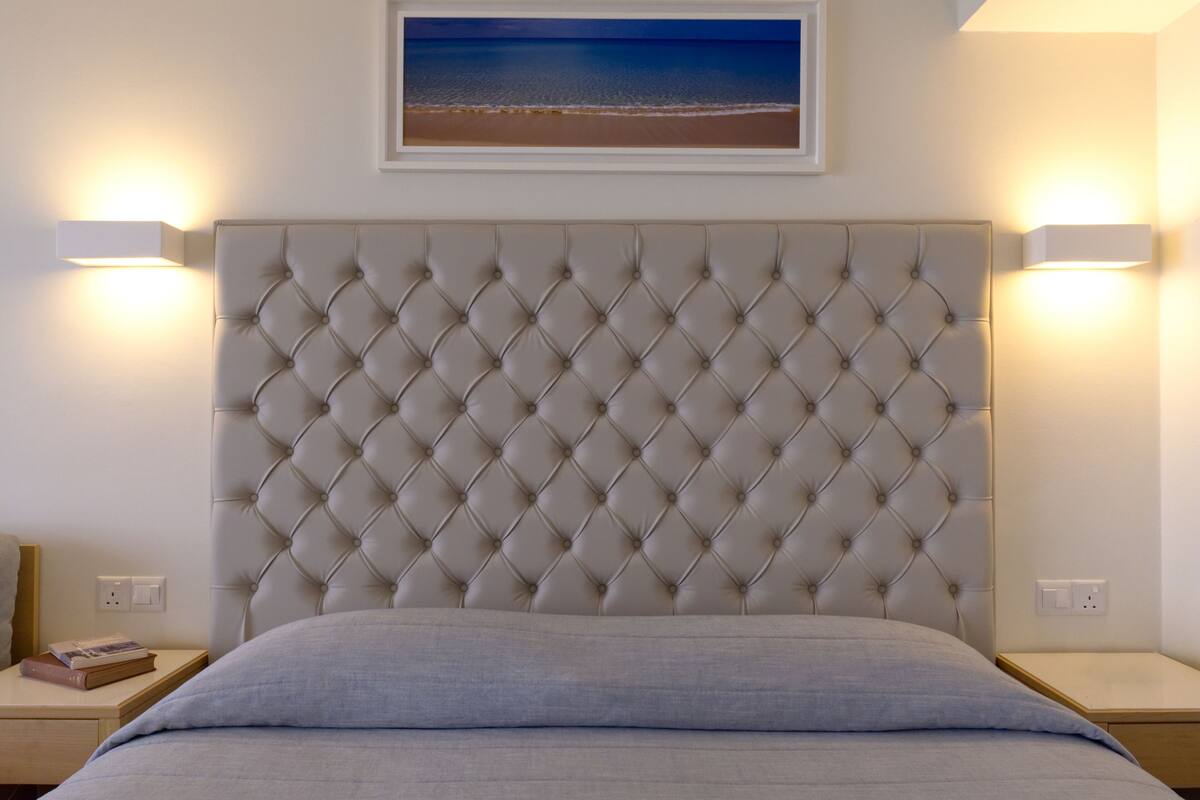 Une chambre à coucher avec une tête de lit capitonnée, des lampes de chevet, une table de chevet et un tableau représentant une plage.