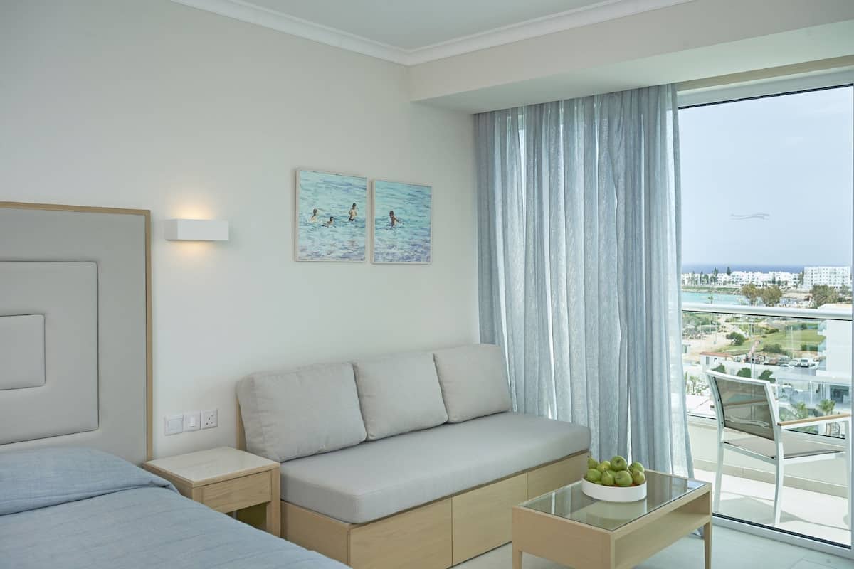 Une chambre d’hôtel moderne avec un canapé, une petite table sur laquelle sont disposés des fruits, un balcon offrant une vue sur la ville et la mer, et des œuvres d’art accrochées au mur.