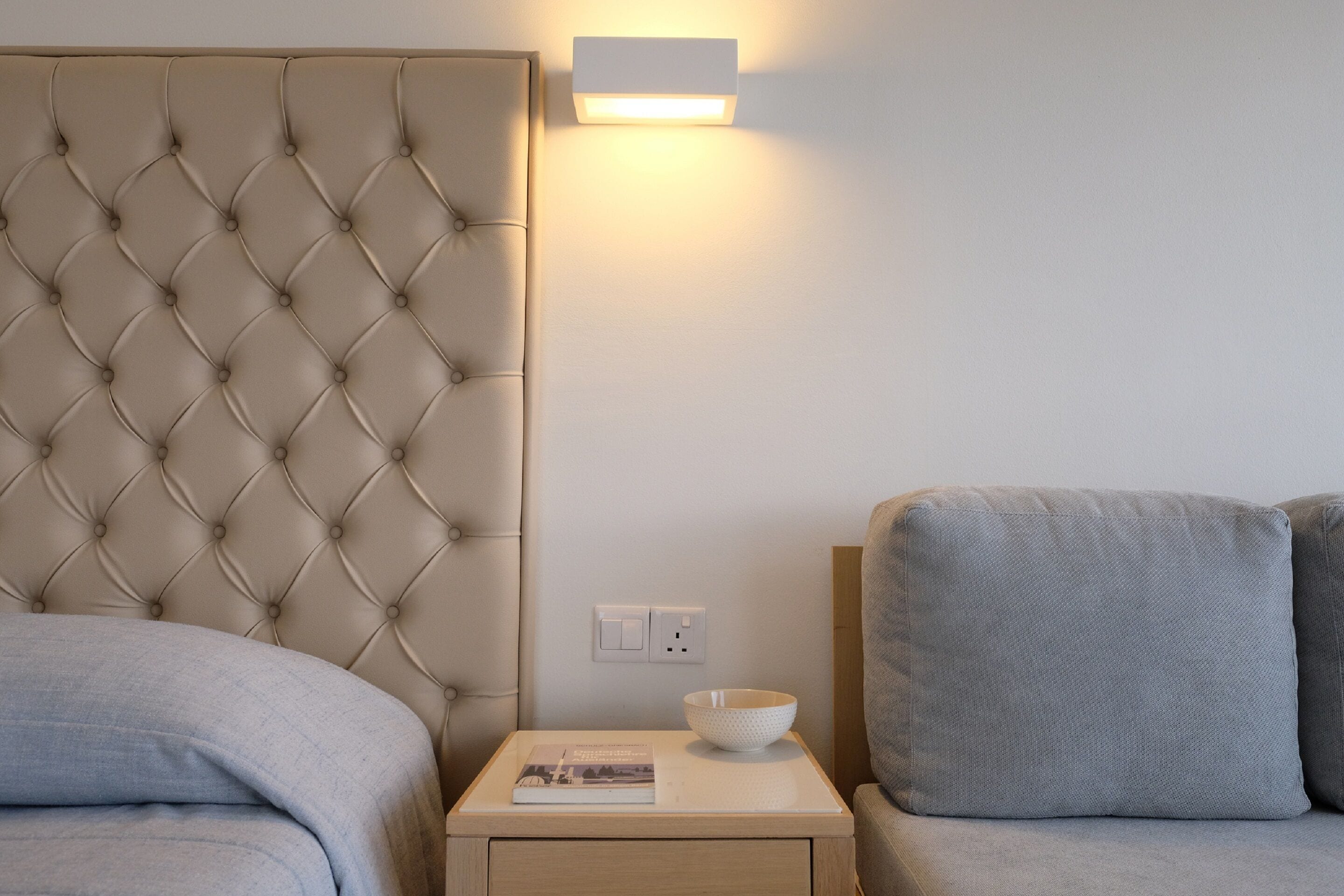 A bedroom with a tufted headboard, a bedside table with a bowl and a book, and a wall-mounted light fixture.