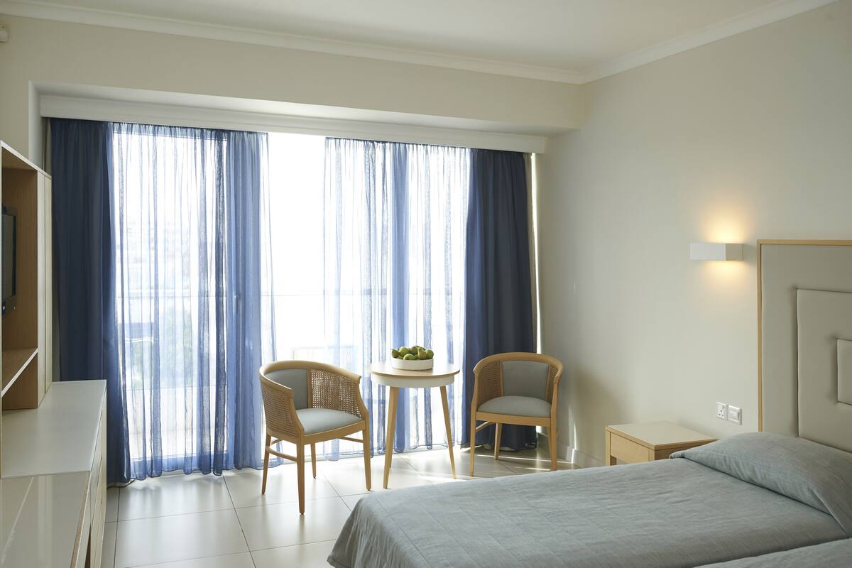 Une chambre d’hôtel avec un lit, deux chaises, une petite table avec une plante et une grande fenêtre avec des rideaux.