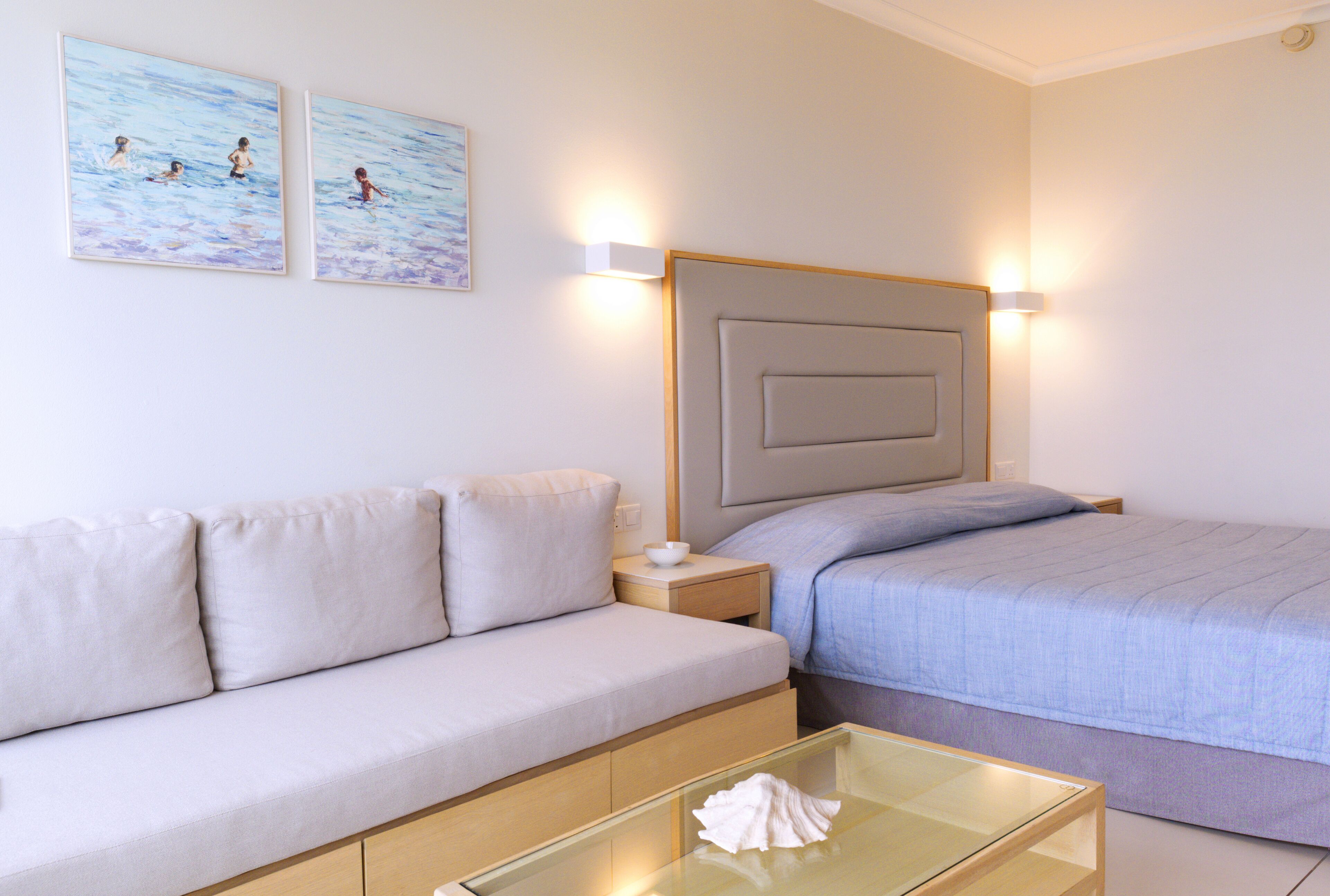 A modern hotel room with a sofa, a bed with a headboard, a bedside table, and two wall-mounted light fixtures.