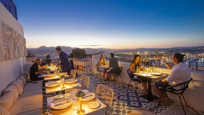 Un espacio exterior para comer con mesas dispuestas para una comida, con vistas a un paisaje urbano al anochecer.