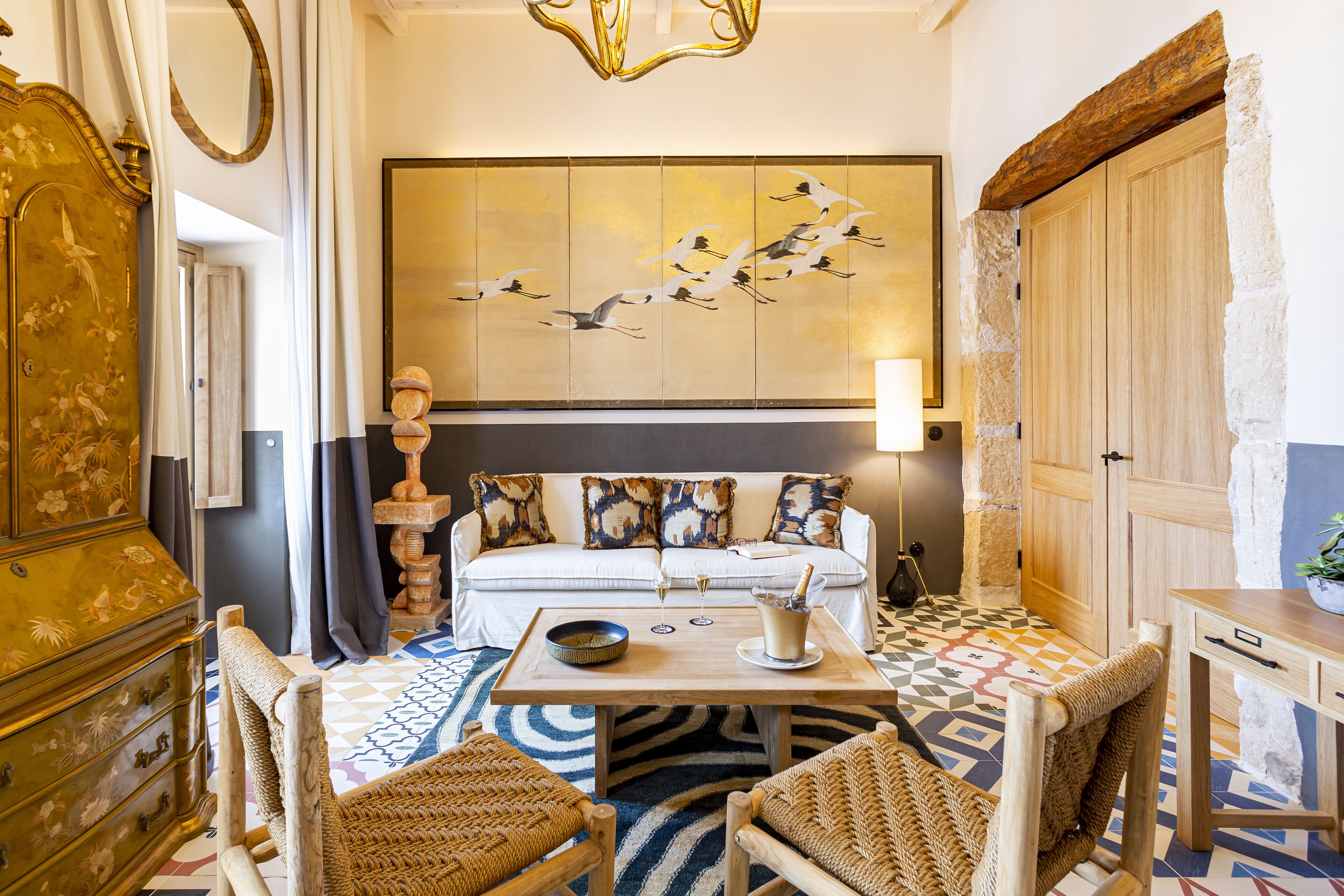 A room with a sofa set, a wooden table, wicker chairs, a decorative wall piece, and a stone wall.