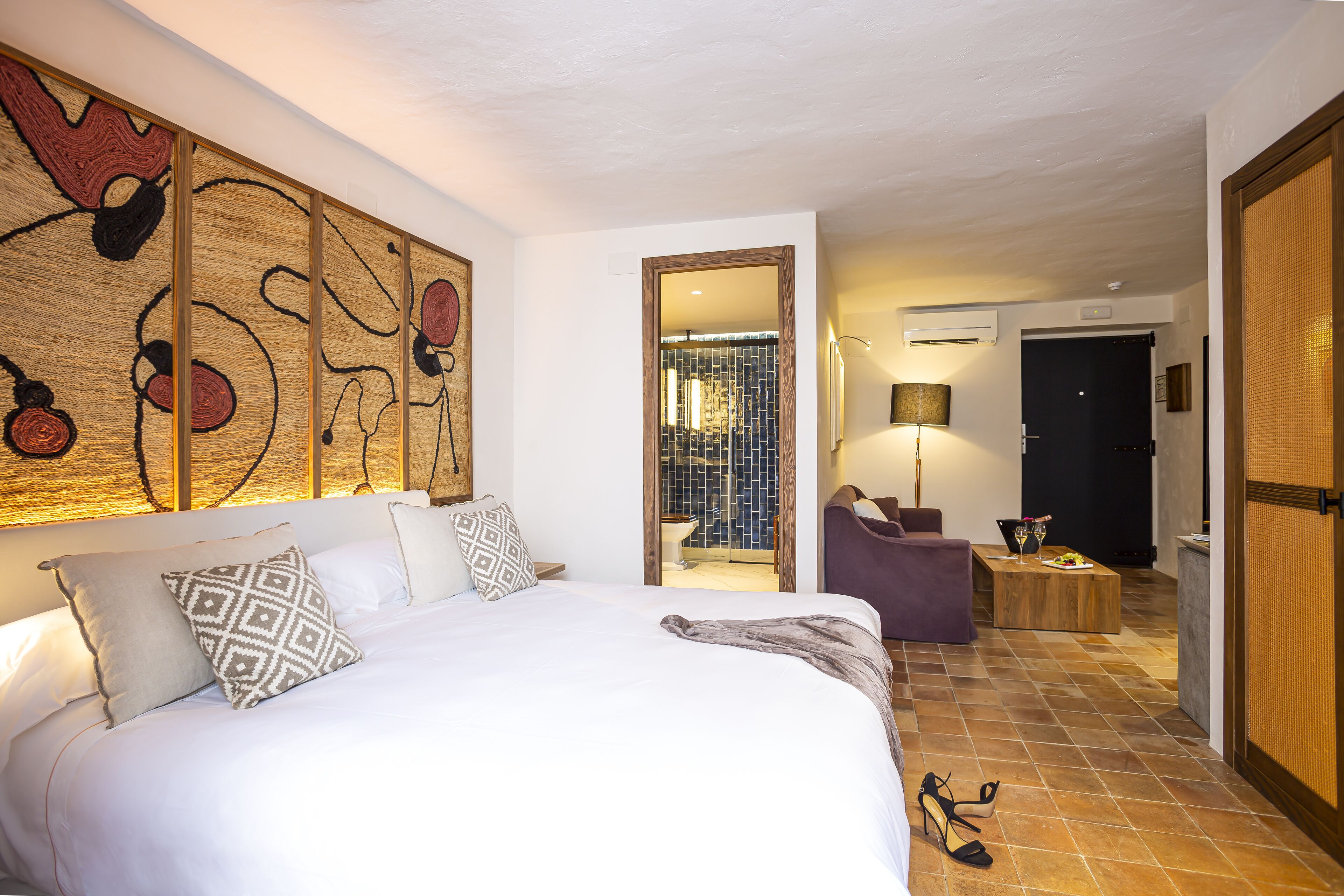 A hotel room with a large bed, a desk, and a chair. There is a door leading to another room.