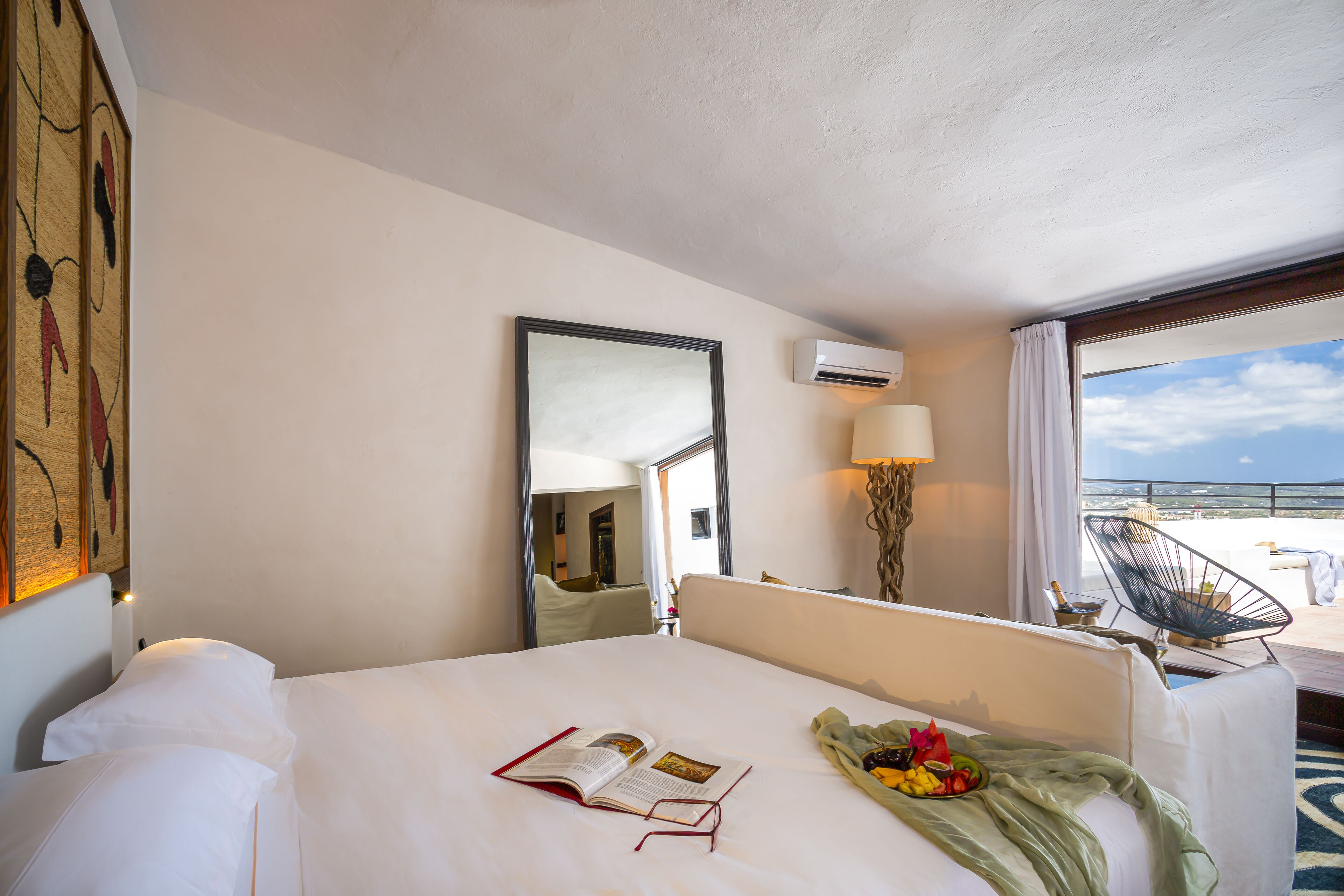 A hotel room with a large bed, a bedside table with a book and a fruit bowl, a chair by the window, and a view of the beach.