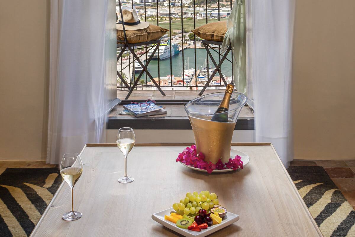 Ein Balkon mit Blick auf einen Hafen, ein Tisch mit Getränken und Obst sowie ein Stuhl.