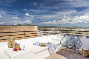A rooftop terrace with a view of a cityscape, a table with a bottle of champagne, a glass, a hat, and a chair.