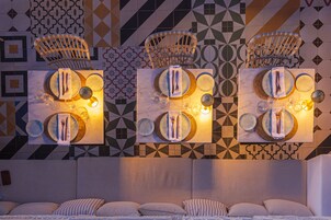 A hotel room with a modern design, featuring a large bed with multiple pillows, a headboard, and a wall-mounted light fixture above a table set with plates, cutlery, and glasses.