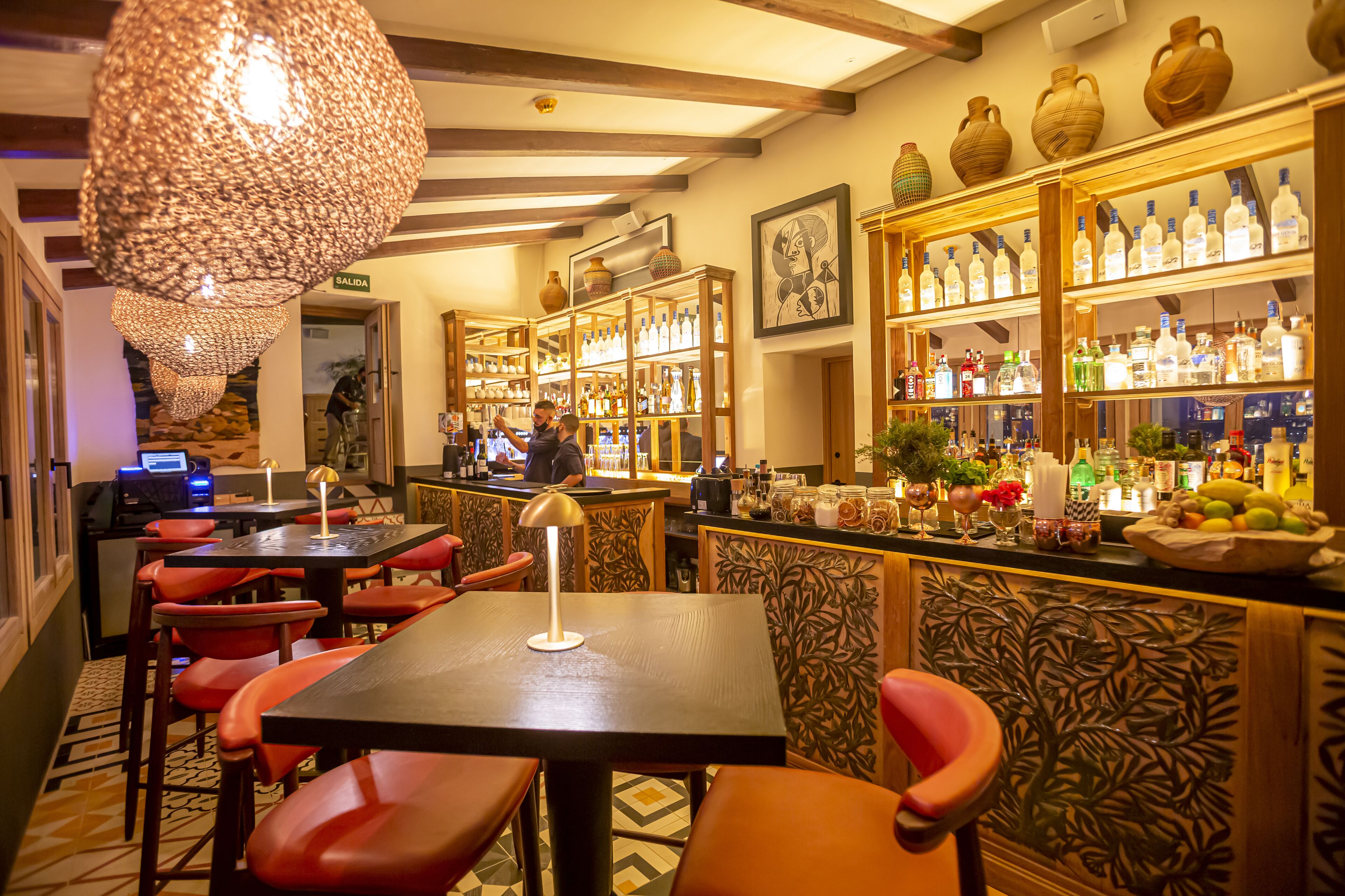 Un área de bar con estantes de madera llenos de botellas, una barra con diversas opciones y un espacio con mesas y sillas.