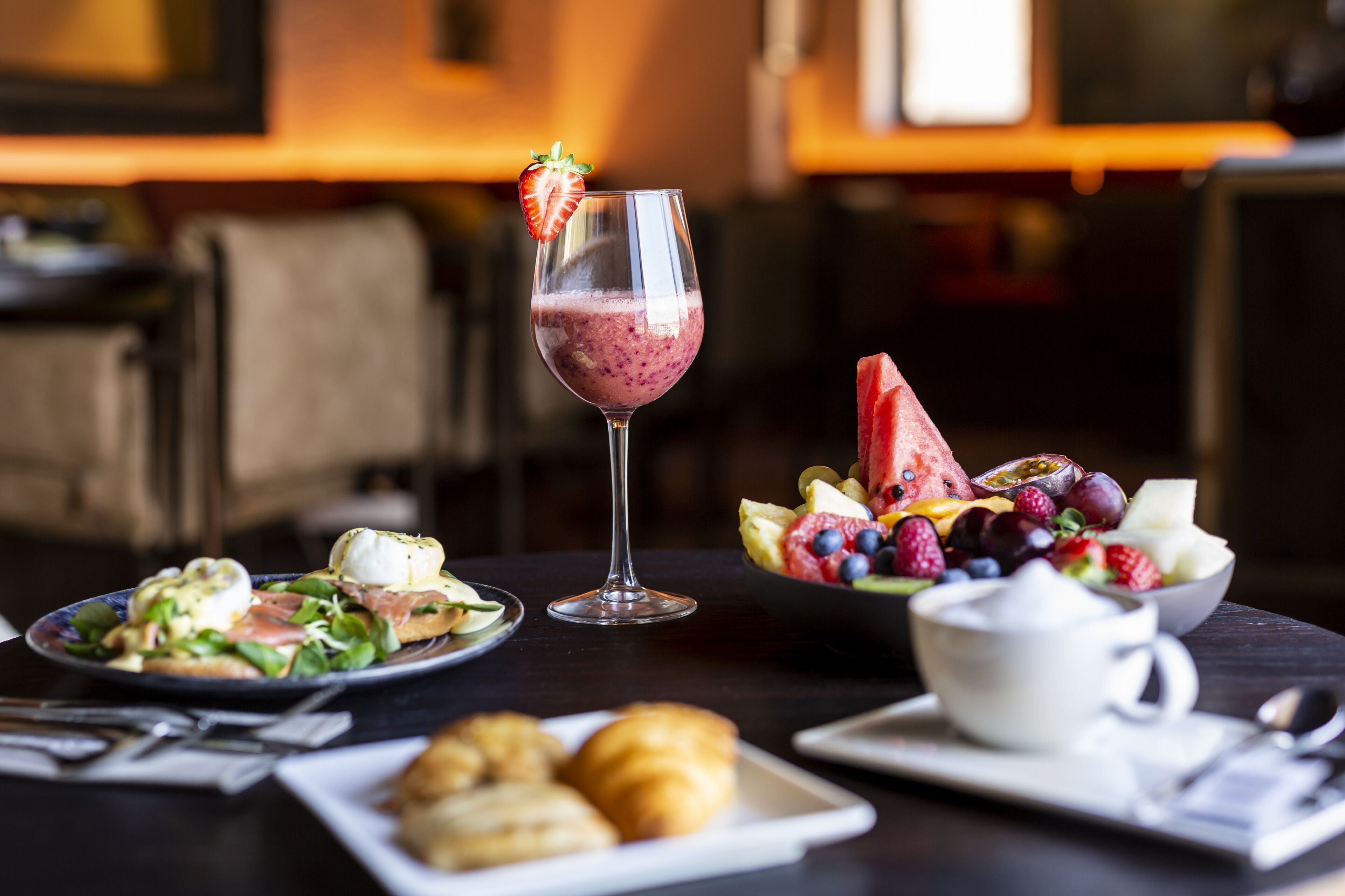 Une table à manger avec une salade, un smoothie dans un verre, un bol de fruits, des croissants et une tasse de café.