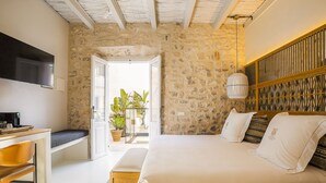 Un dormitorio con pared de piedra, cama con ropa blanca, cabecero de madera, televisor, escritorio con silla y planta en maceta.