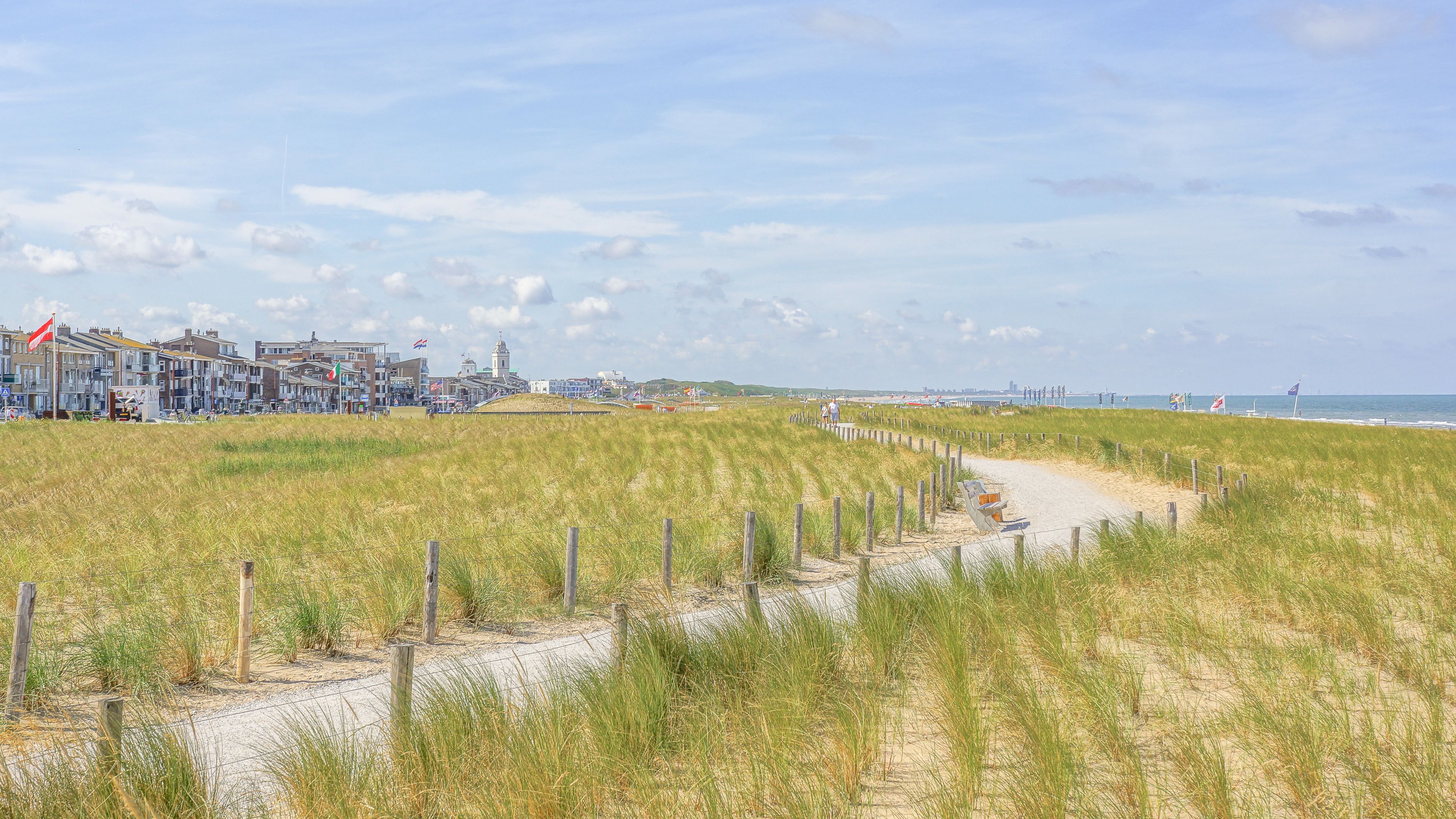 Un sentier côtier, avec un chemin de sable menant à une plage, bordé de dunes herbeuses et d’une rangée de bâtiments ornés de drapeaux.