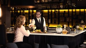 Zona de bar con un cantinero sirviendo una bebida a un cliente. Se observan estantes con botellas y copas al fondo.